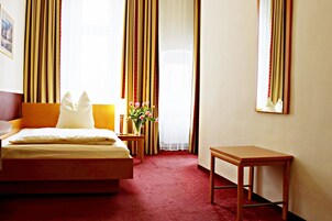 Habitación de hotel con cama, mesita de noche con lámpara, una pequeña mesa y ventana con cortinas.