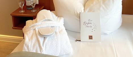 A neatly made bed with white linens, a bottle of water, two glasses, a robe, and a card on the bedside table.