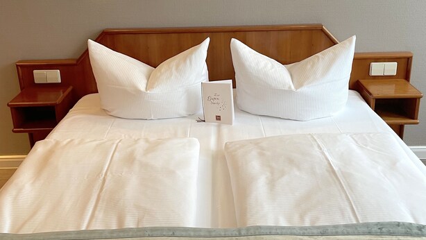 Una cama bien hecha con sábanas y almohadas blancas, cabecera de madera y dos mesitas de noche con lámparas.