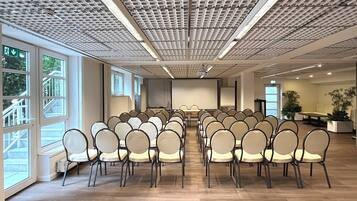 A conference room with rows of chairs facing a central table, large windows on one side, and a ceiling with a grid pattern.