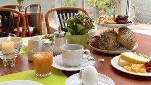 Mesa de café da manhã com pratos, xícaras, um candeeiro, um vaso de flores e diversos pães e frutas.