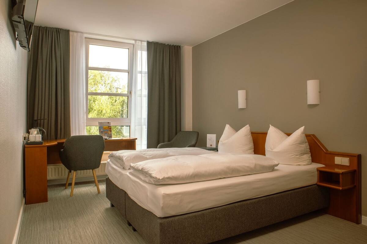 Una habitación de hotel con una cama grande, un escritorio, una silla y un ventanal con cortinas.