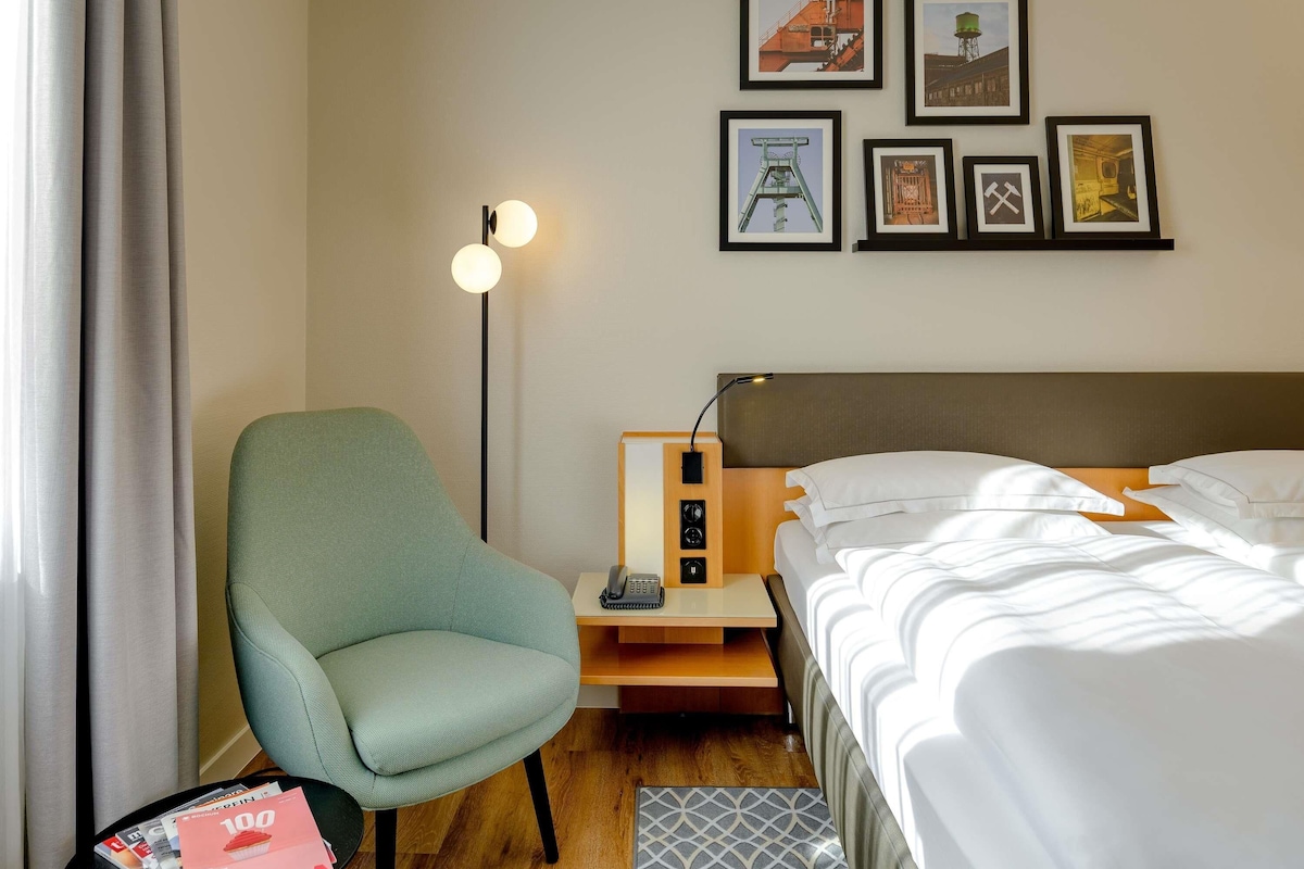Una habitación de hotel con cama, una silla, un escritorio con lámpara y cuadros en la pared.