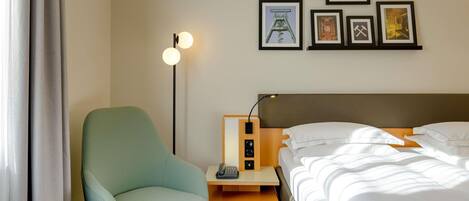 A hotel room with a bed, a chair, a desk with a lamp, and framed pictures on the wall.