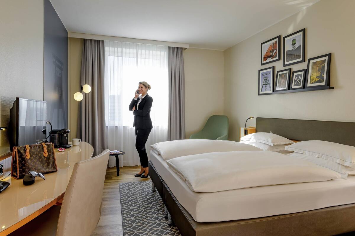 Une chambre d’hôtel comprenant un lit, un bureau avec un ordinateur, une chaise, une machine à café et une fenêtre avec des rideaux.