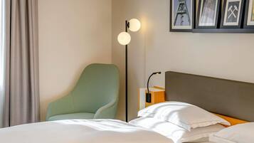 A hotel room with a bed, a green armchair, a floor lamp, and framed pictures on the wall.