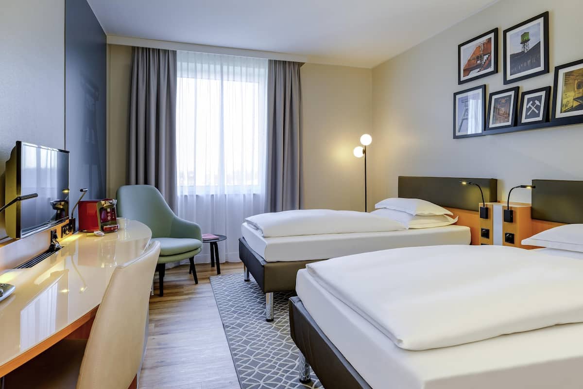 Une chambre d’hôtel avec deux lits, un bureau avec une machine à café, une chaise, un lampadaire et des tableaux encadrés au mur.