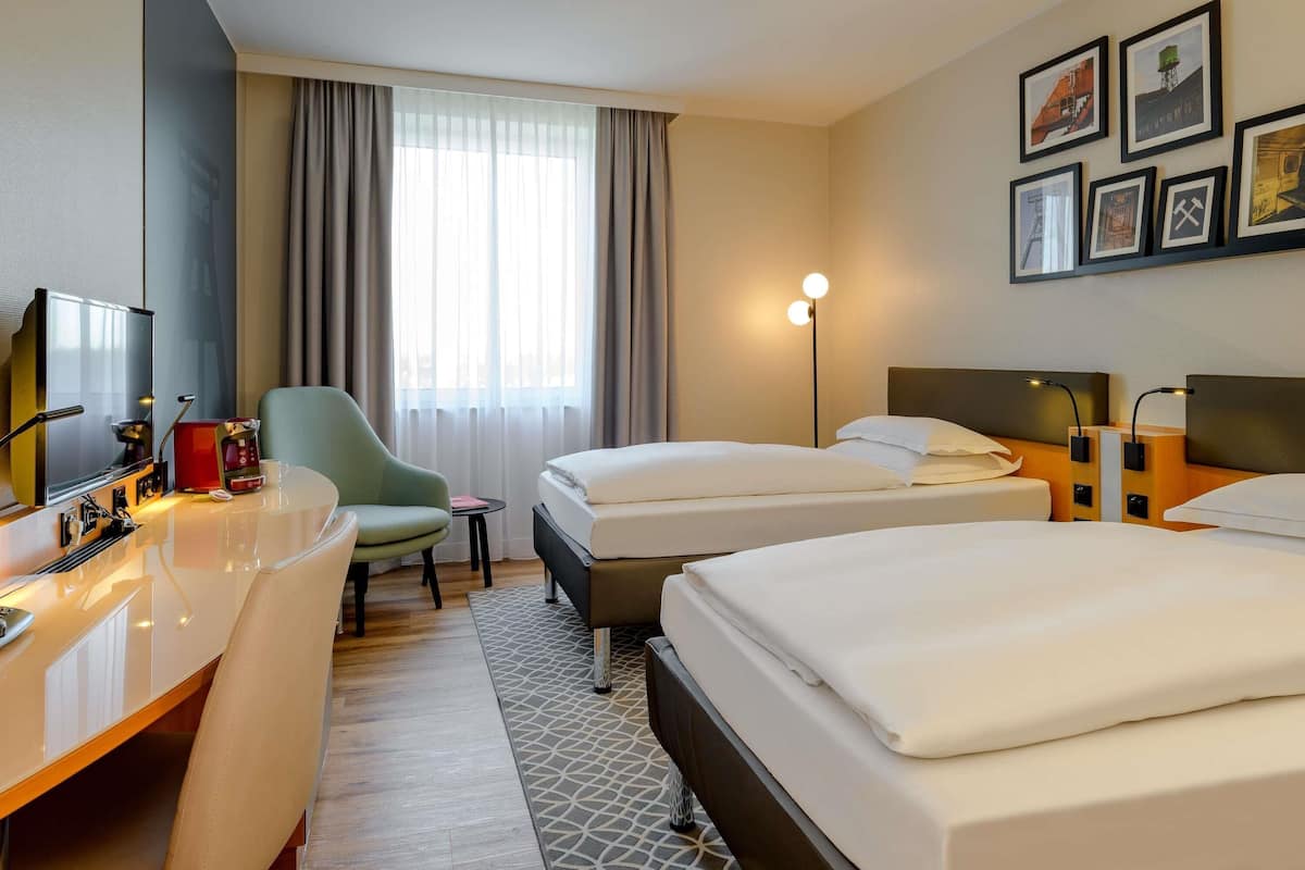 A hotel room with a desk, two beds, a chair, a small table, a lamp, and framed pictures on the wall.