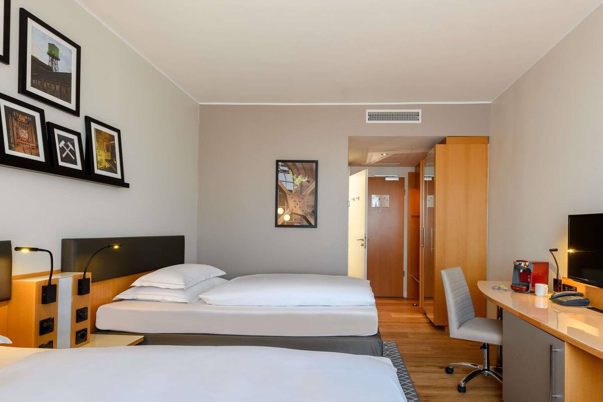 A hotel room with two beds, a desk with a computer, a TV, and framed pictures on the wall.