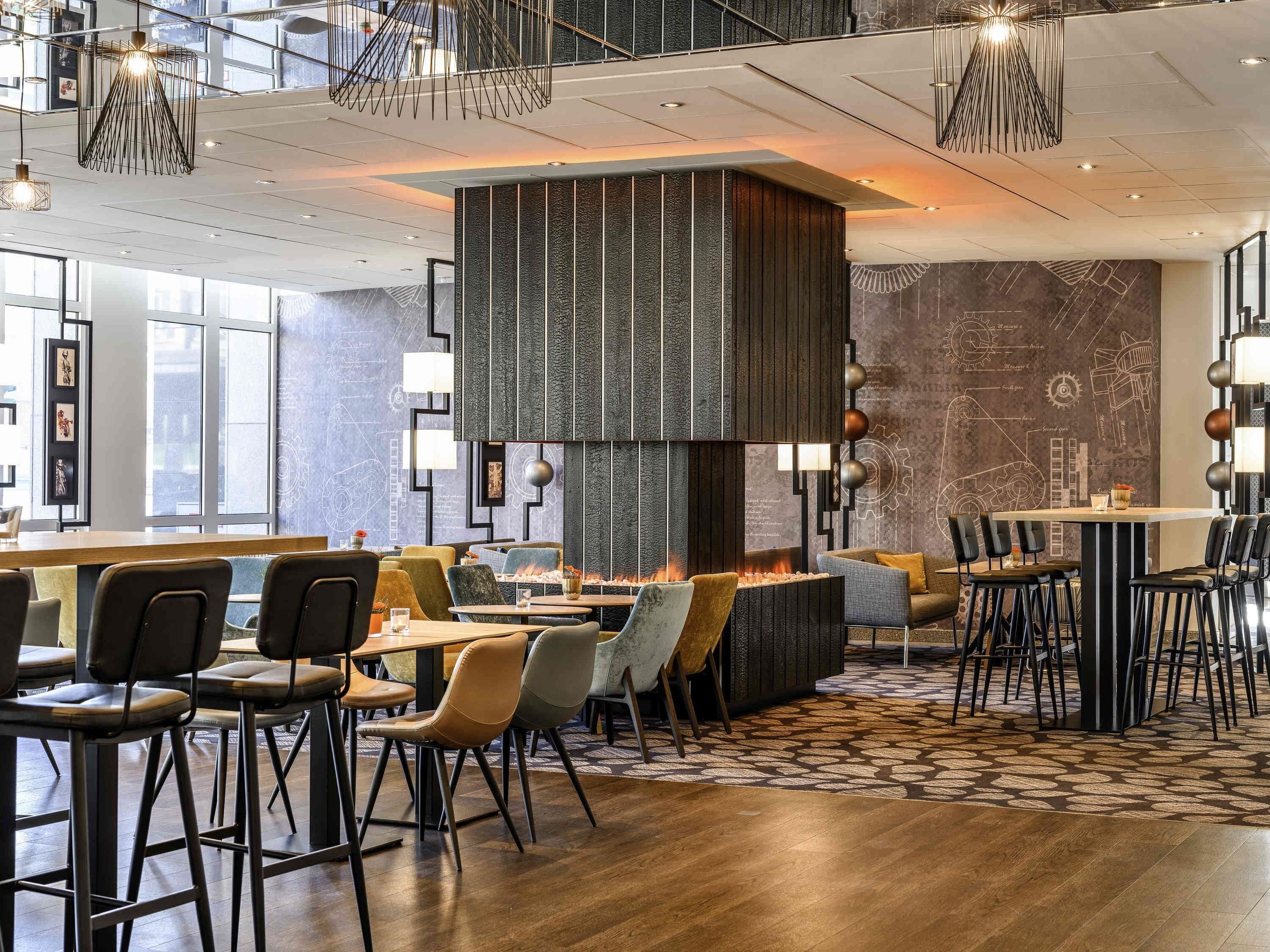 A modern restaurant with high ceilings, pendant lighting, and a central fire feature.