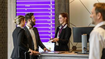 Eine Rezeption mit digitalem Display, das Namen und Symbole anzeigt, ein modernes Interieur mit hölzernem Empfangstresen und einer schlichten, geometrischen Leuchte.