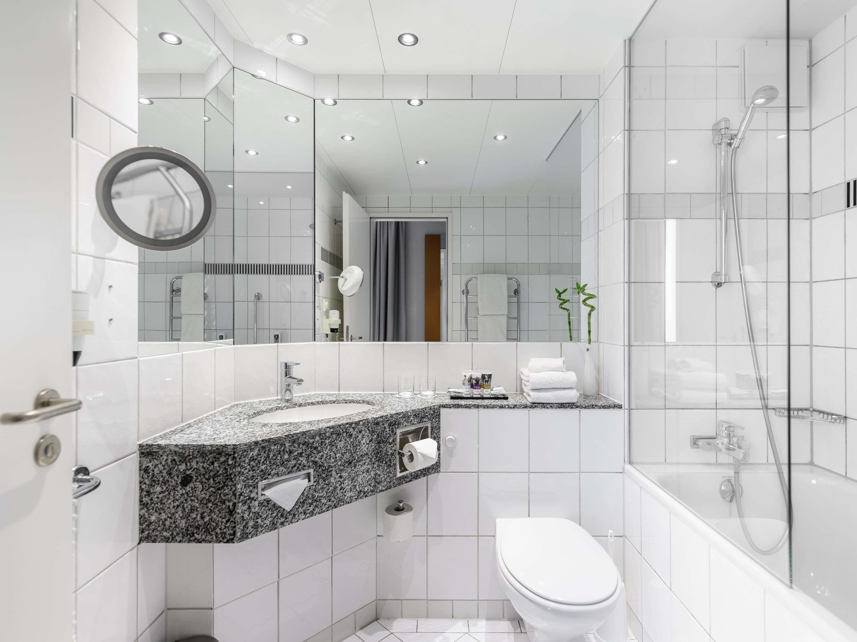 Un baño moderno con encimera de granito, inodoro blanco, ducha con mampara de vidrio y un espejo grande.