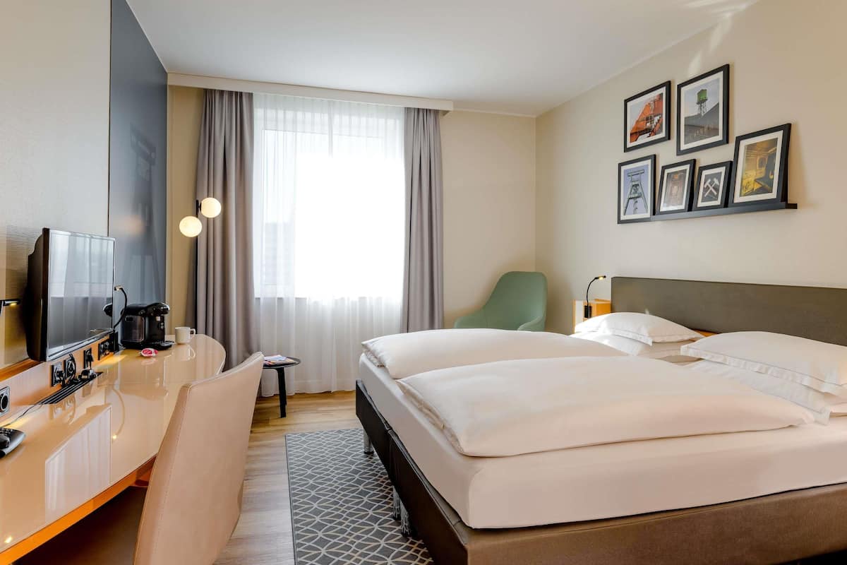 A hotel room with a bed, a desk with a coffee maker, a chair, a TV, and framed pictures on the wall.