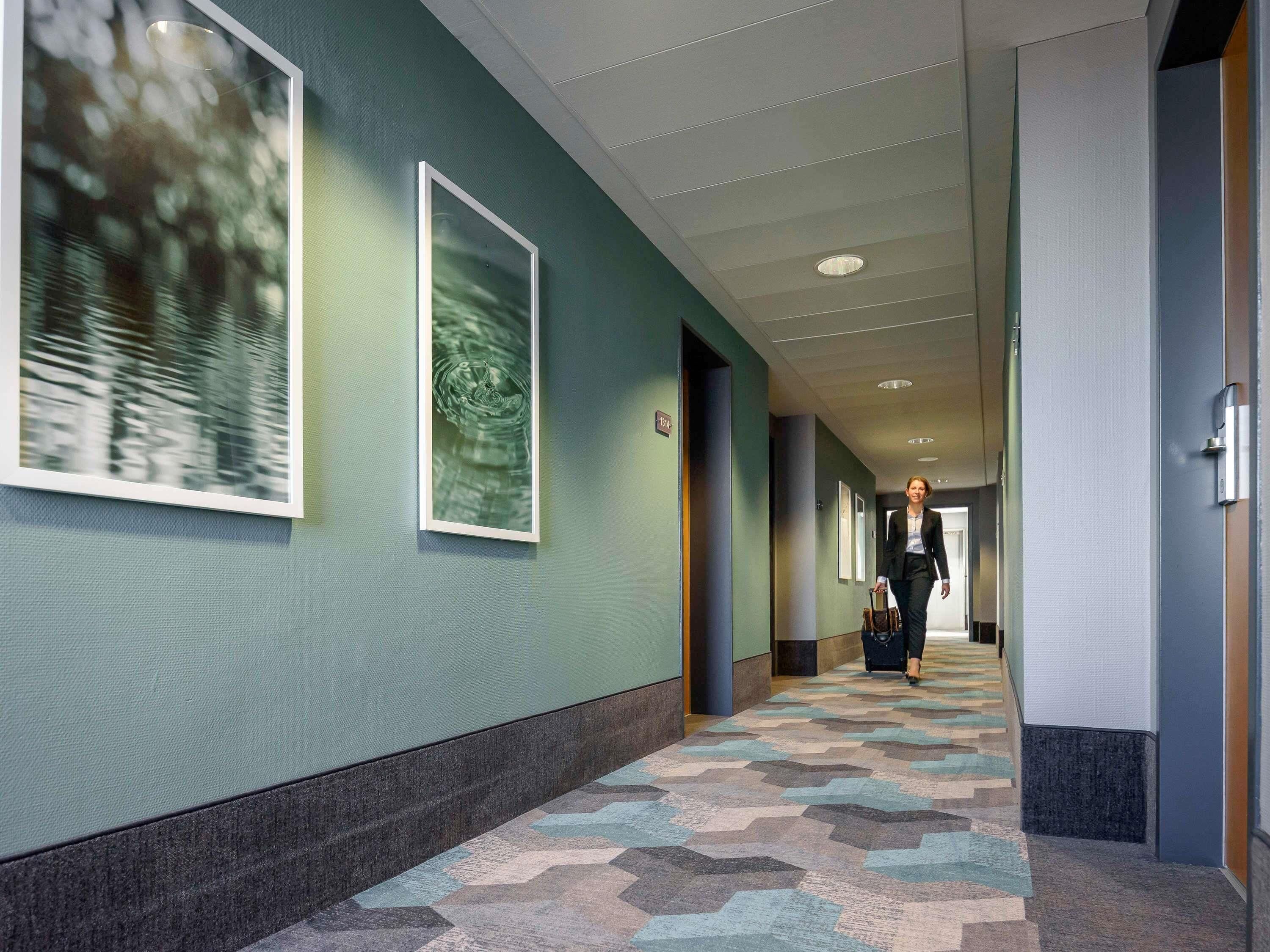A hotel corridor with framed artwork on the walls, a patterned carpet, and a person walking towards the end of the hallway.