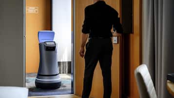 A man in a black shirt and pants standing in a hotel room, facing a robotic vacuum cleaner.