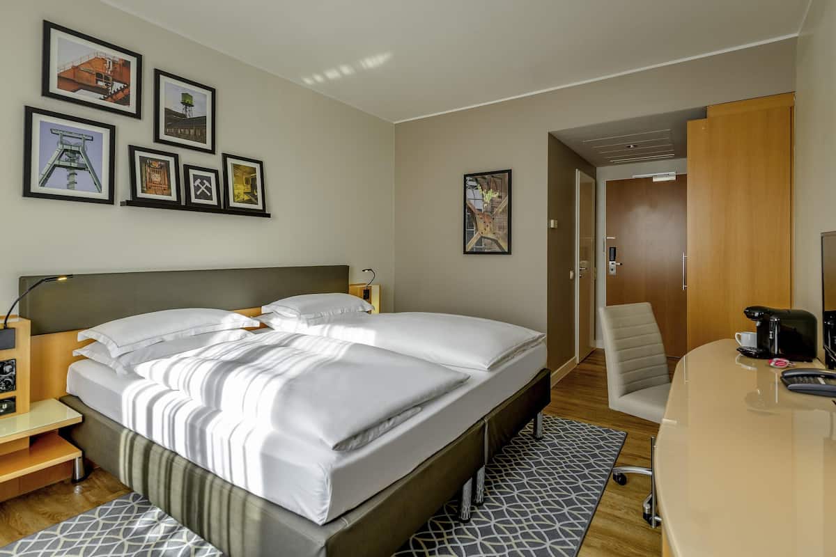 A hotel room with a bed, bedside tables, a desk with a chair, a coffee maker, and framed pictures on the wall.