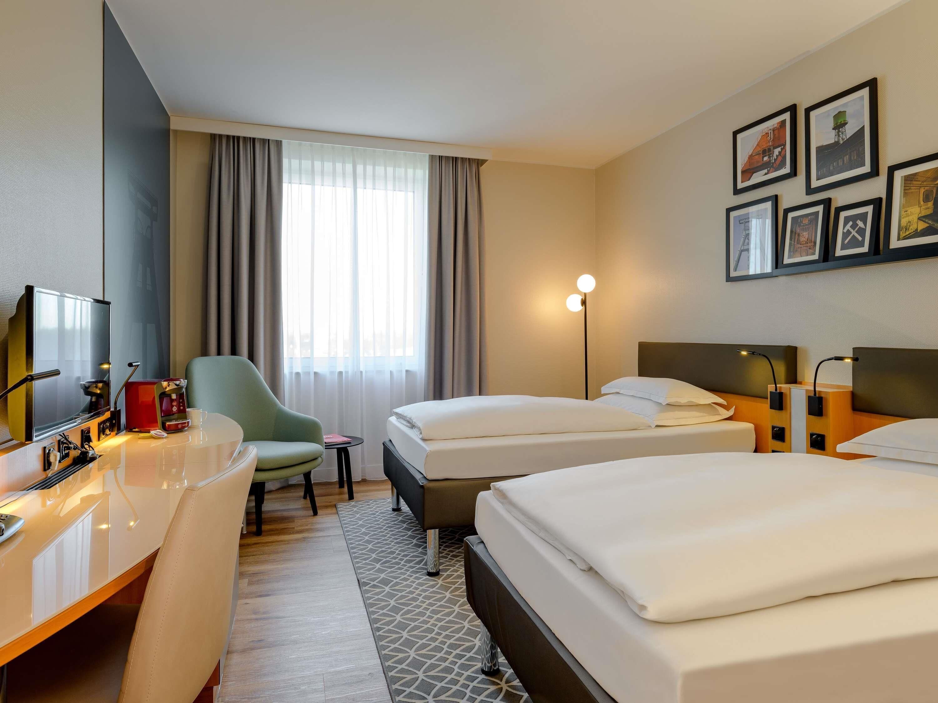 Une chambre d’hôtel équipée d’un bureau, de deux lits, d’une chaise, d’une petite table, d’une lampe et de tableaux encadrés accrochés au mur.