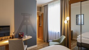 A hotel room with a bed, a desk with a lamp, a chair, a coffee machine, and a TV.