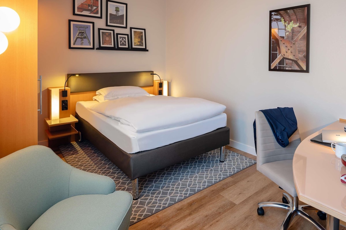 A hotel room with a bed, bedside tables, a desk, a chair, and a wall with framed pictures.