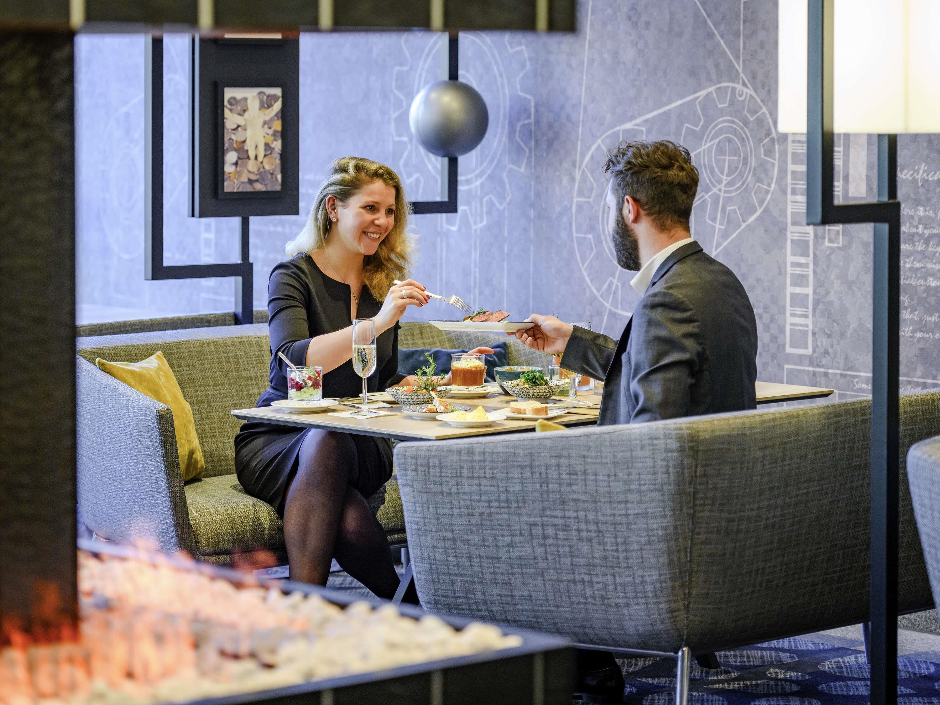 Un coin repas avec une table dressée pour deux personnes, un canapé et un mur décoratif orné de motifs abstraits.