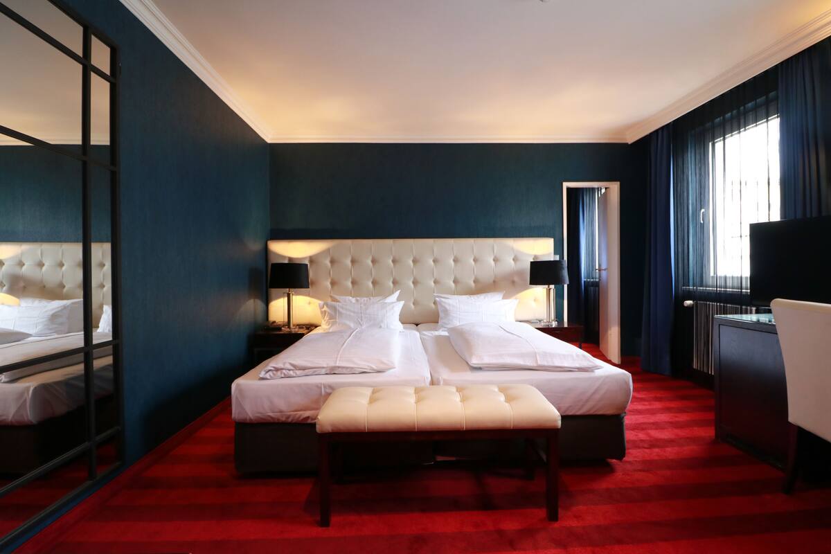 A hotel room with a large bed, a bench, a desk, and a mirror.