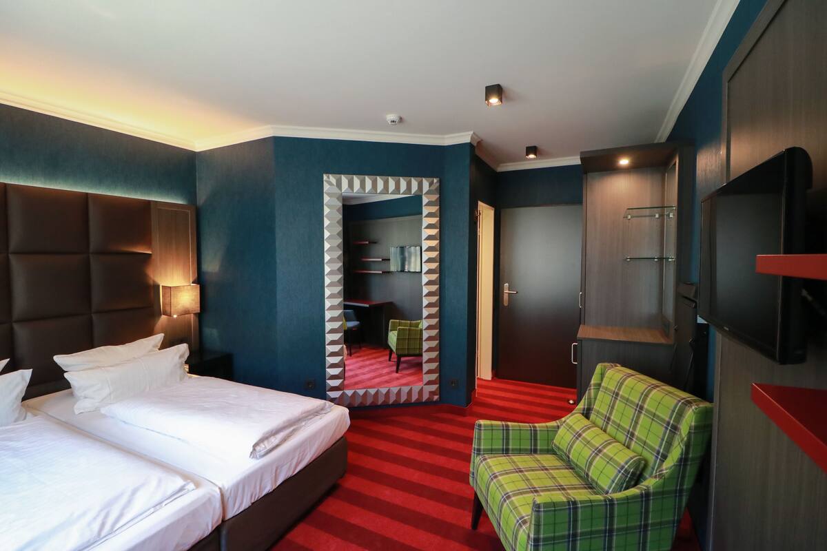 A hotel room with a large bed, a green plaid chair, a TV, and a mirror with a geometric frame.