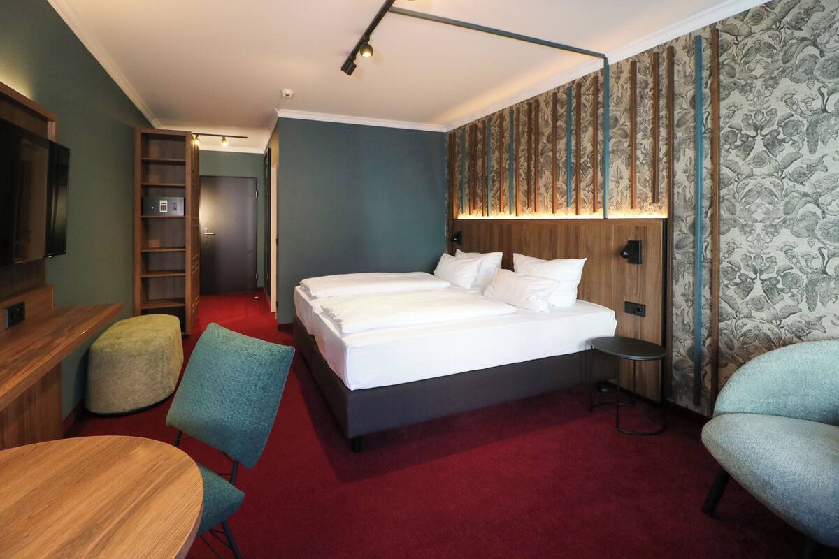 A hotel room with a large bed, a desk, a chair, a small table, a wardrobe, and a TV.