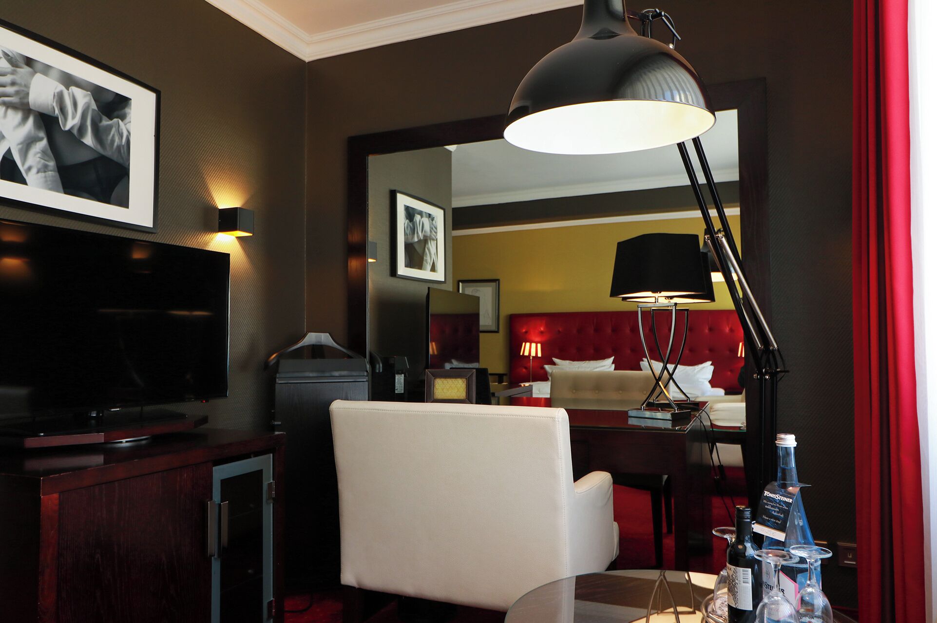 A hotel room with a television, a desk with a lamp, a chair, and a table with bottles.