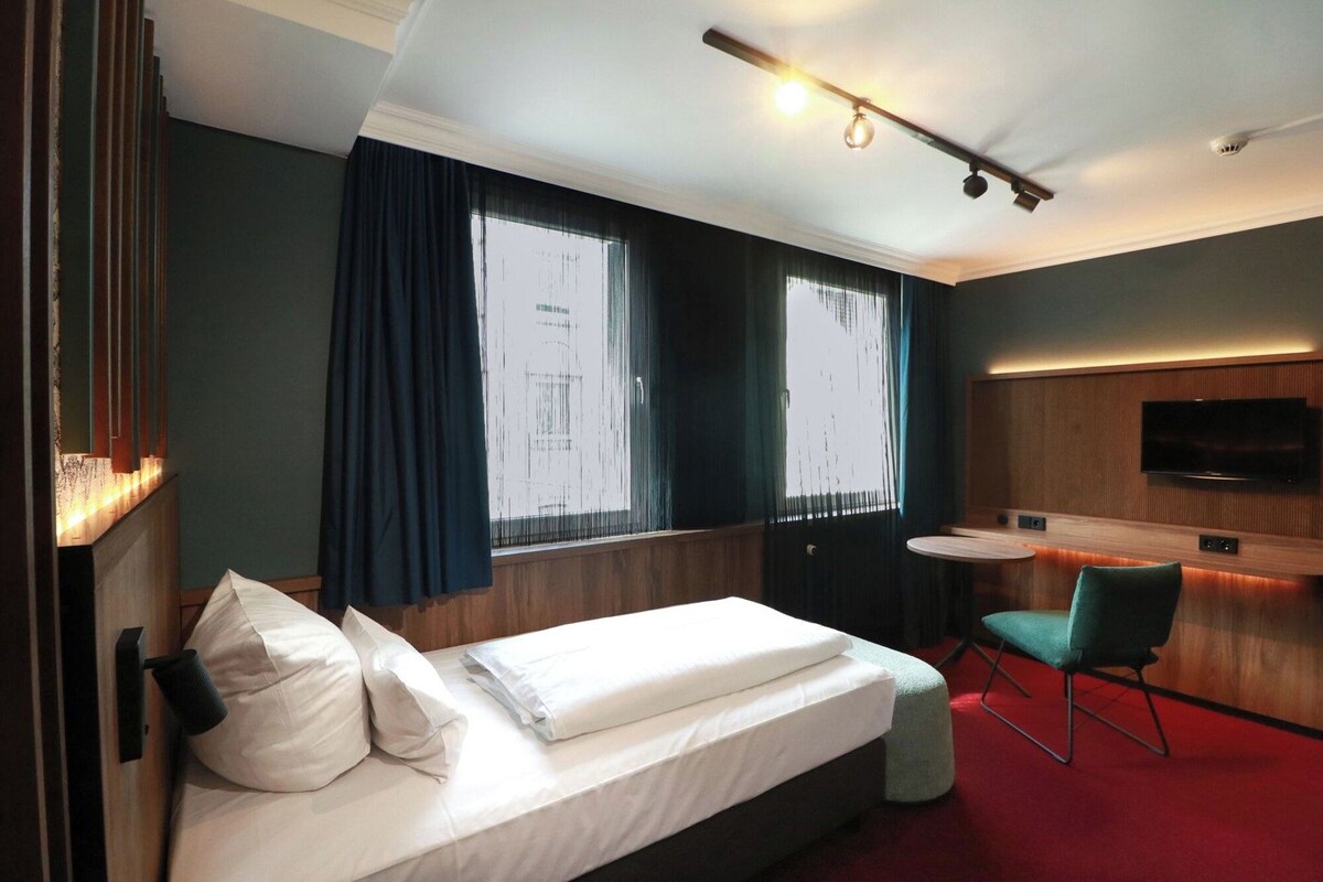 Habitación de hotel con cama, escritorio, silla, televisión y ventana con cortinas.