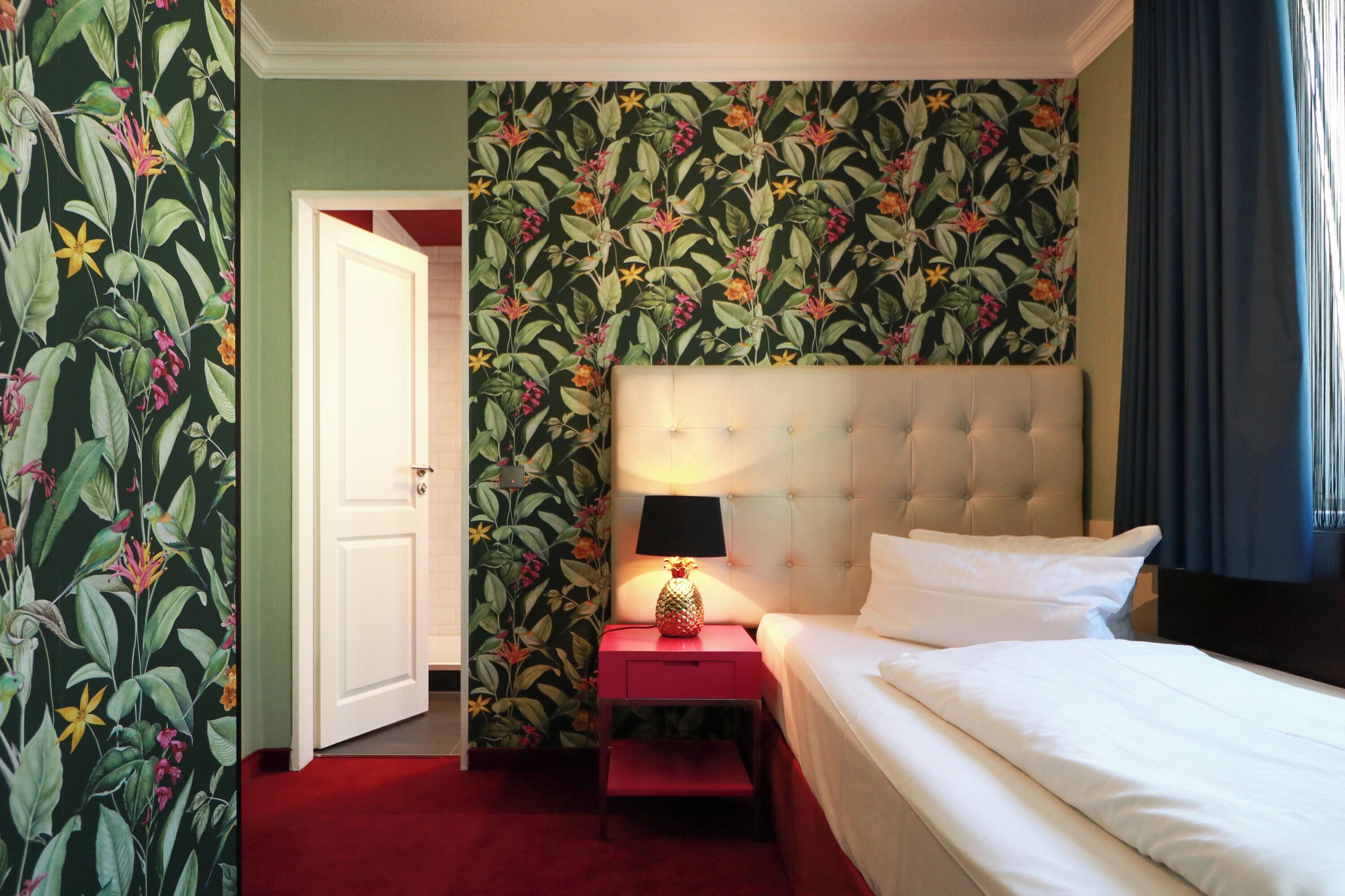 Habitación de hotel con papel tapiz floral, cama con ropa de cama blanca, mesita de noche roja con lámpara y puerta que da a otra habitación.