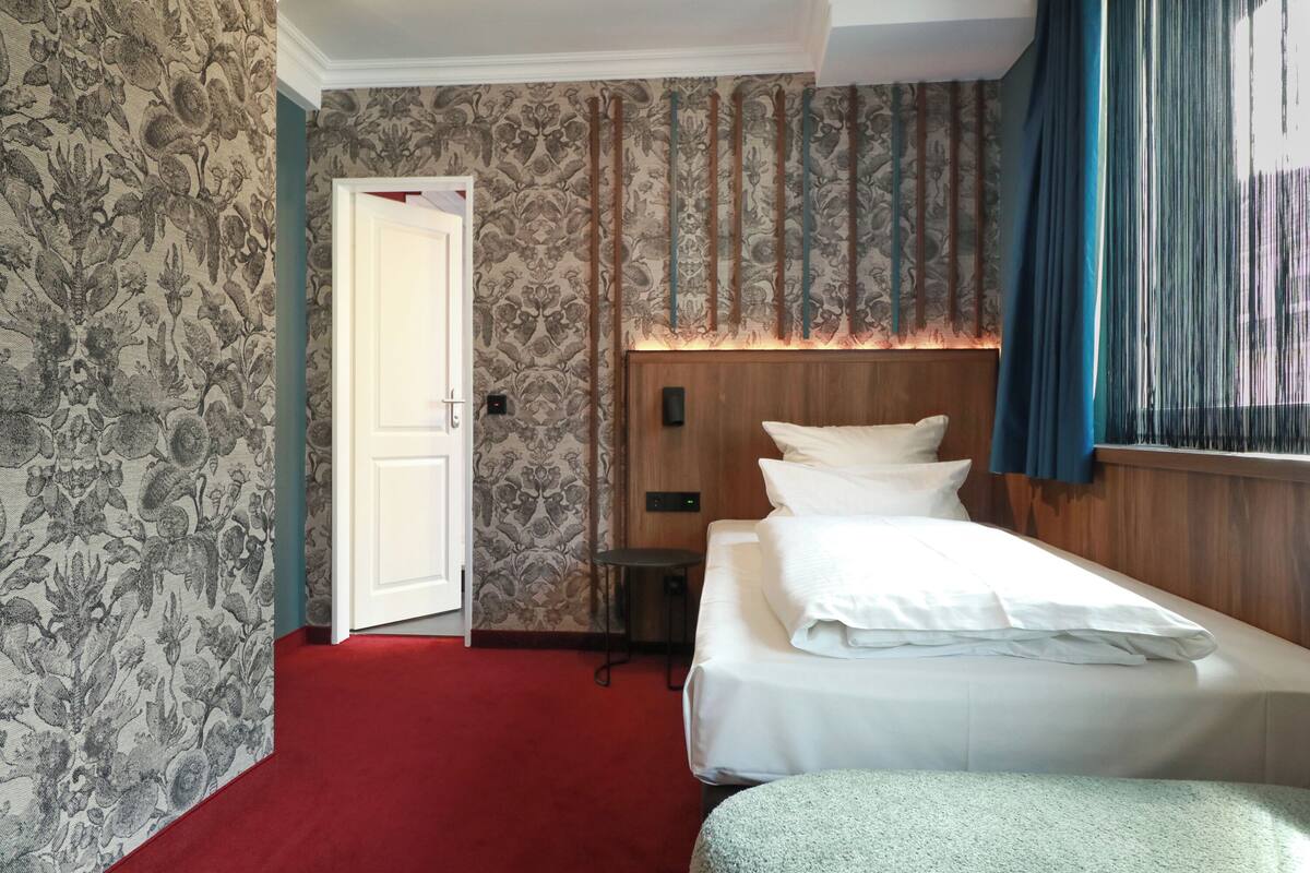 A hotel room with a bed, a small table, a wall with vertical blinds, and a door.