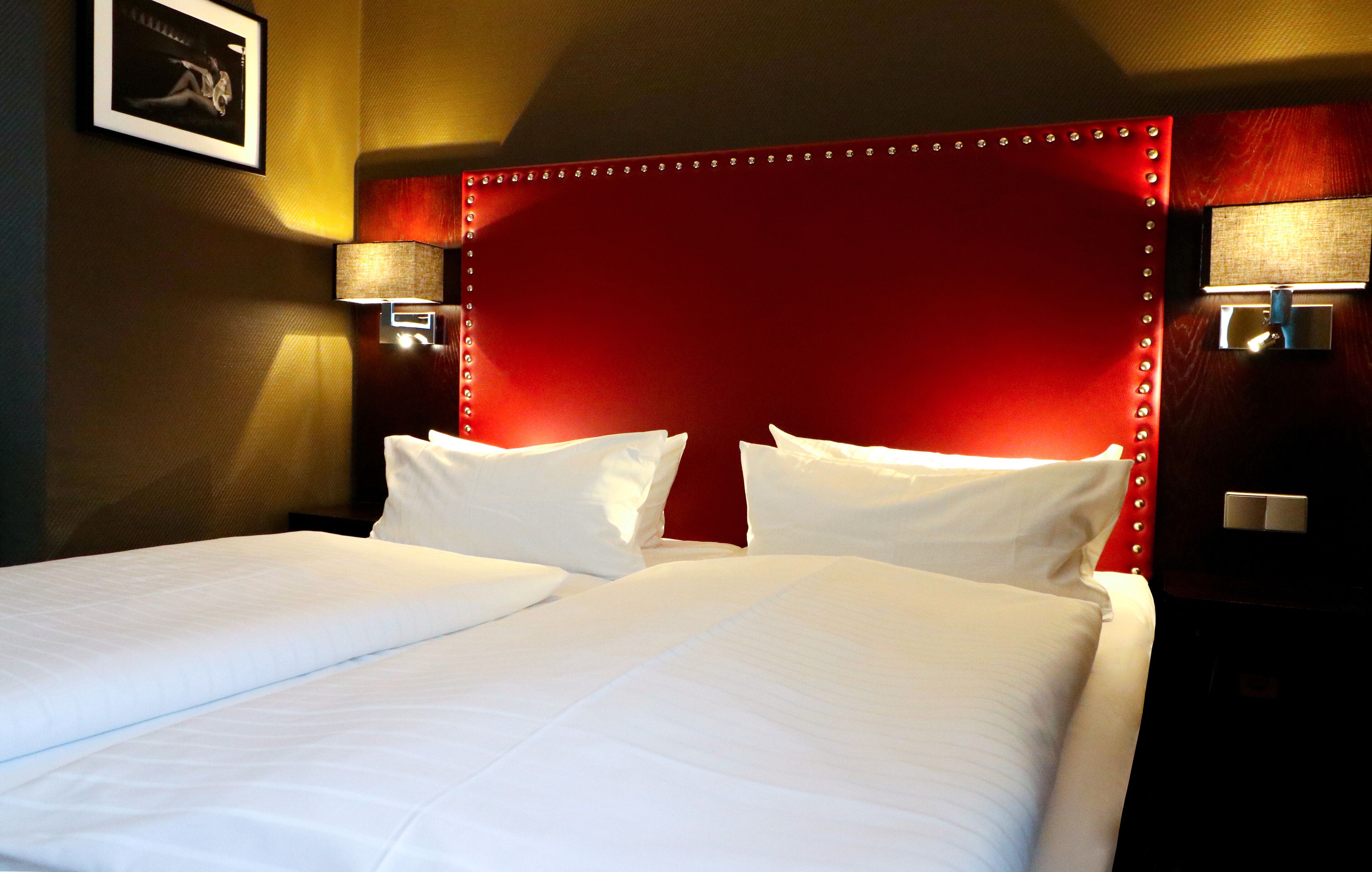 A double bed with white linens, a red upholstered headboard, and two wall-mounted lamps on either side.