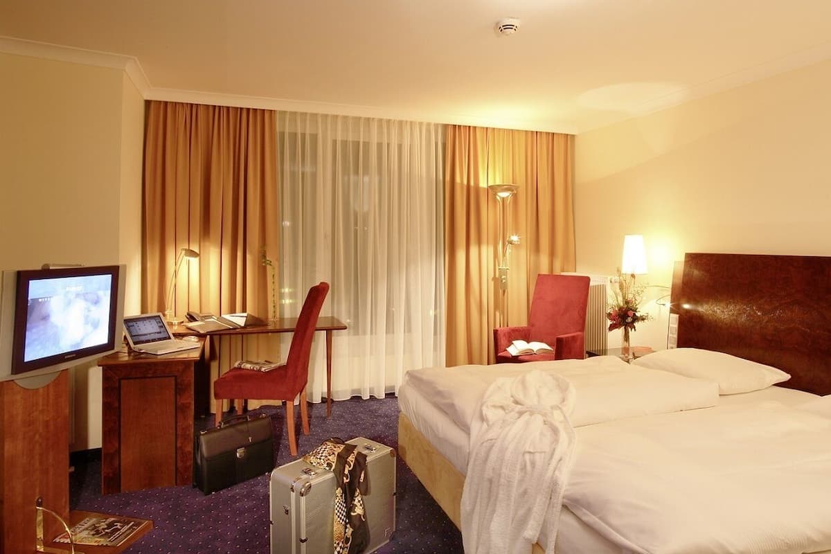 A hotel room with a bed, a desk with a laptop, a chair, a TV, and a suitcase.