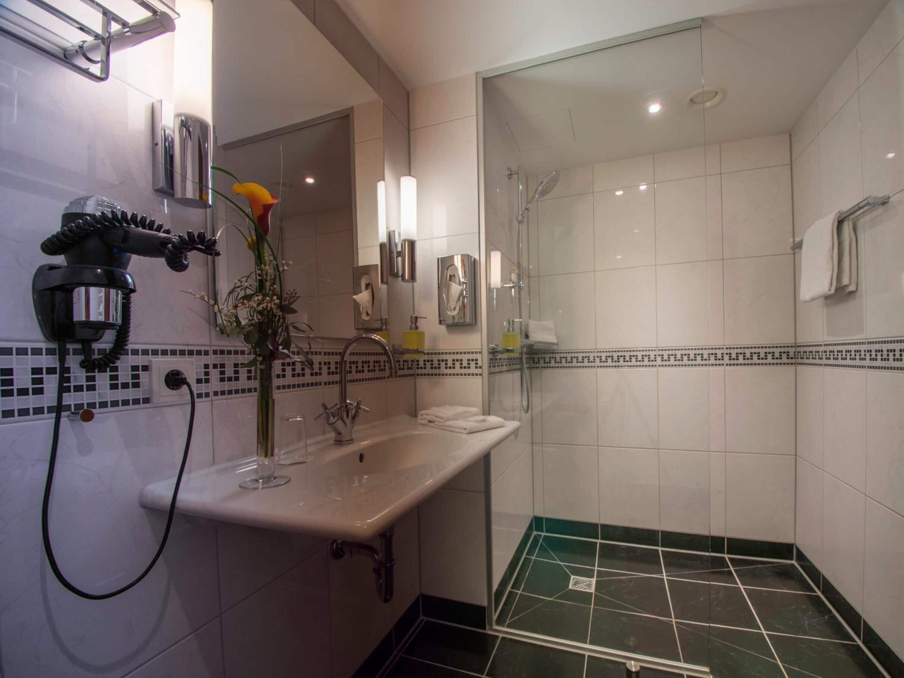 A modern bathroom with a glass shower enclosure, a sink with a vase of flowers, and a hairdryer mounted on the wall.