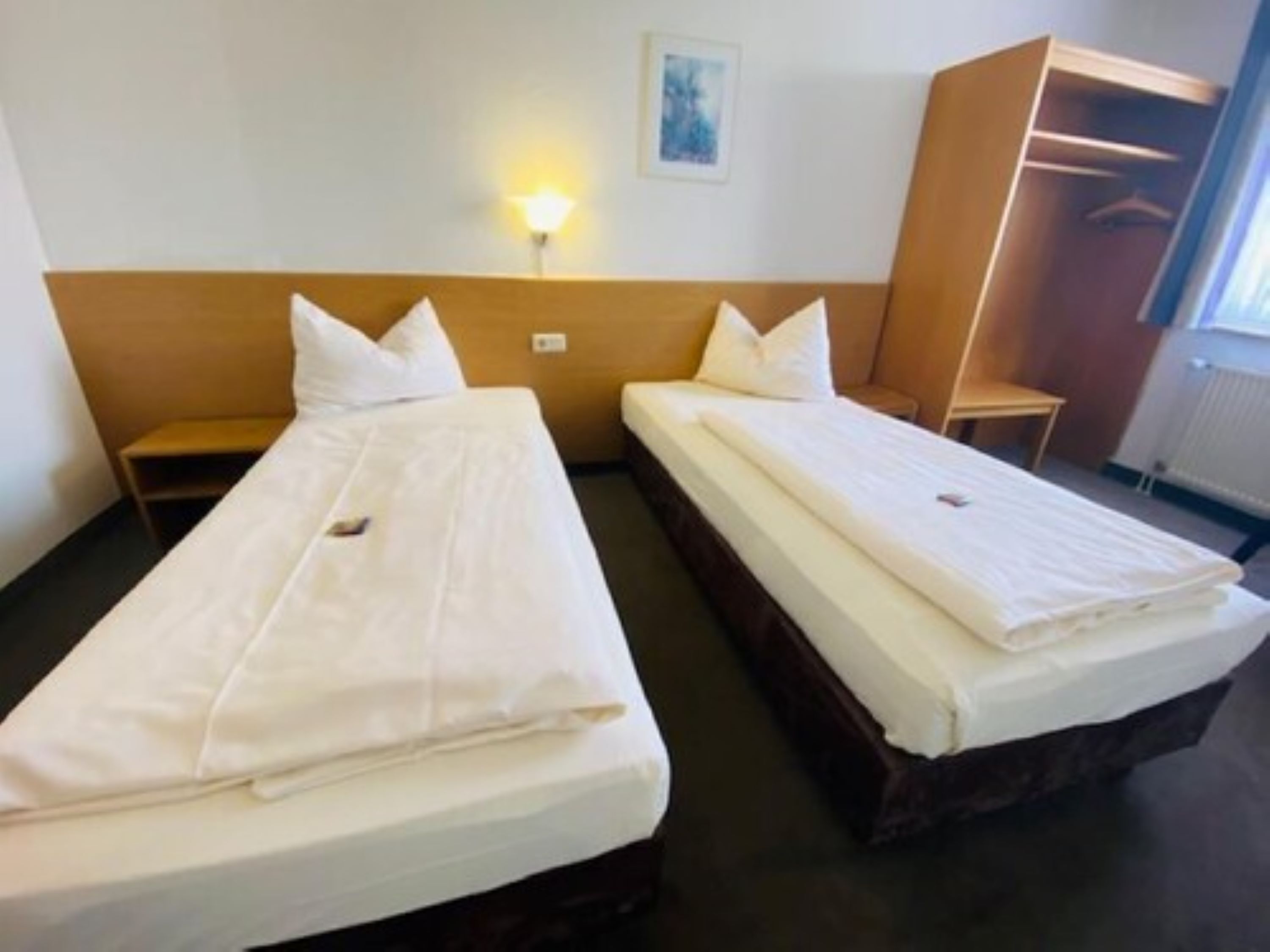 A hotel room with two single beds, a nightstand, a wardrobe, and a wall-mounted light.