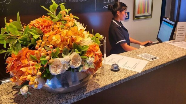A hotel reception desk with a large floral arrangement, a chalkboard with messages, and a computer monitor. - Hotel Christina (Cologne)