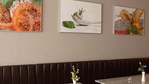 A restaurant interior with a long dark brown booth seating area, multiple tables with white marble tops, and framed artwork on the wall. - Hotel Christina (Cologne)