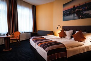 Un cuarto de hotel con una cama grande, un escritorio con silla, una ventana con cortinas y un cuadro de paisaje urbano en la pared.