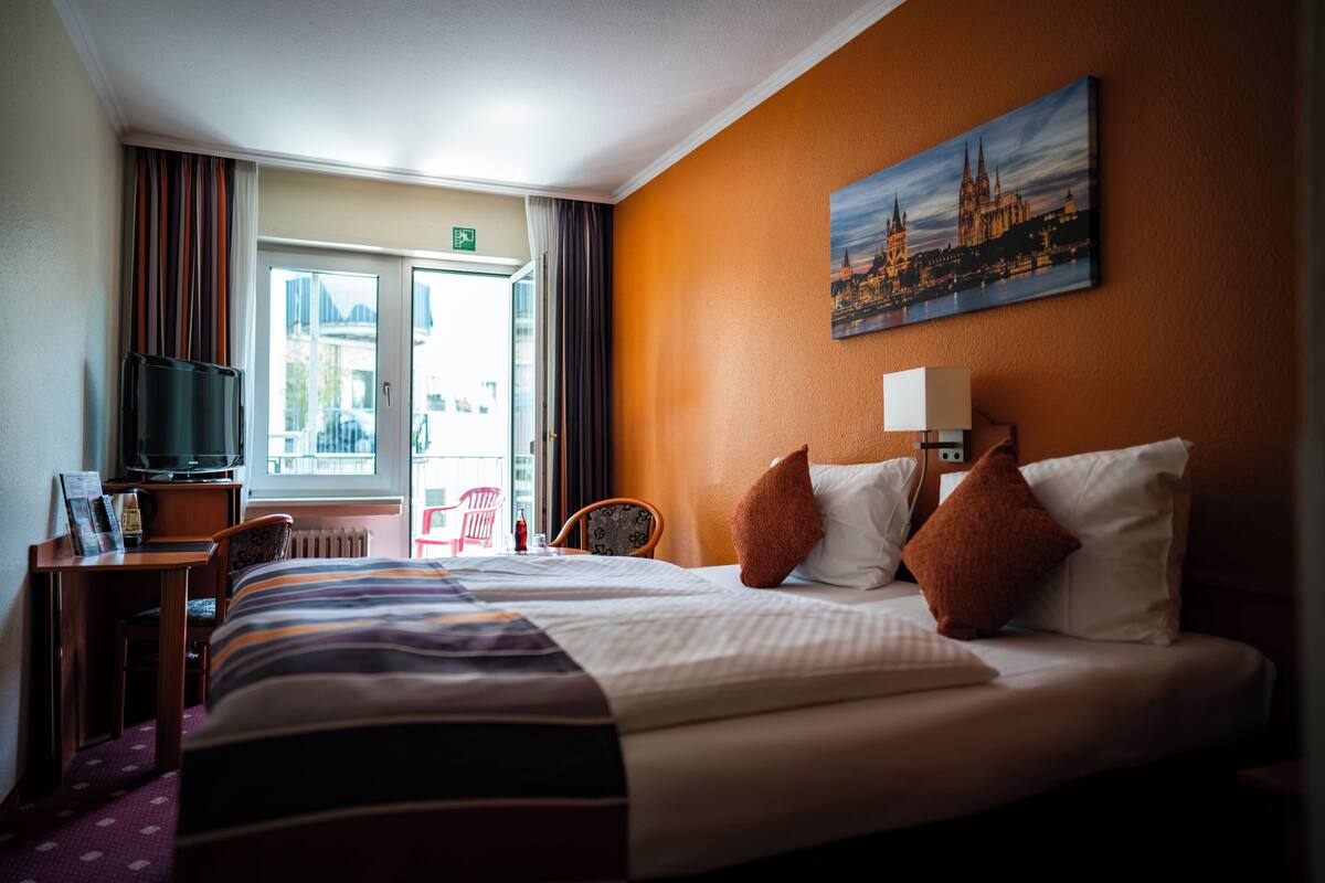 Habitación de hotel con una cama grande, un escritorio con televisor, una silla y un cuadro de un paisaje urbano en la pared.