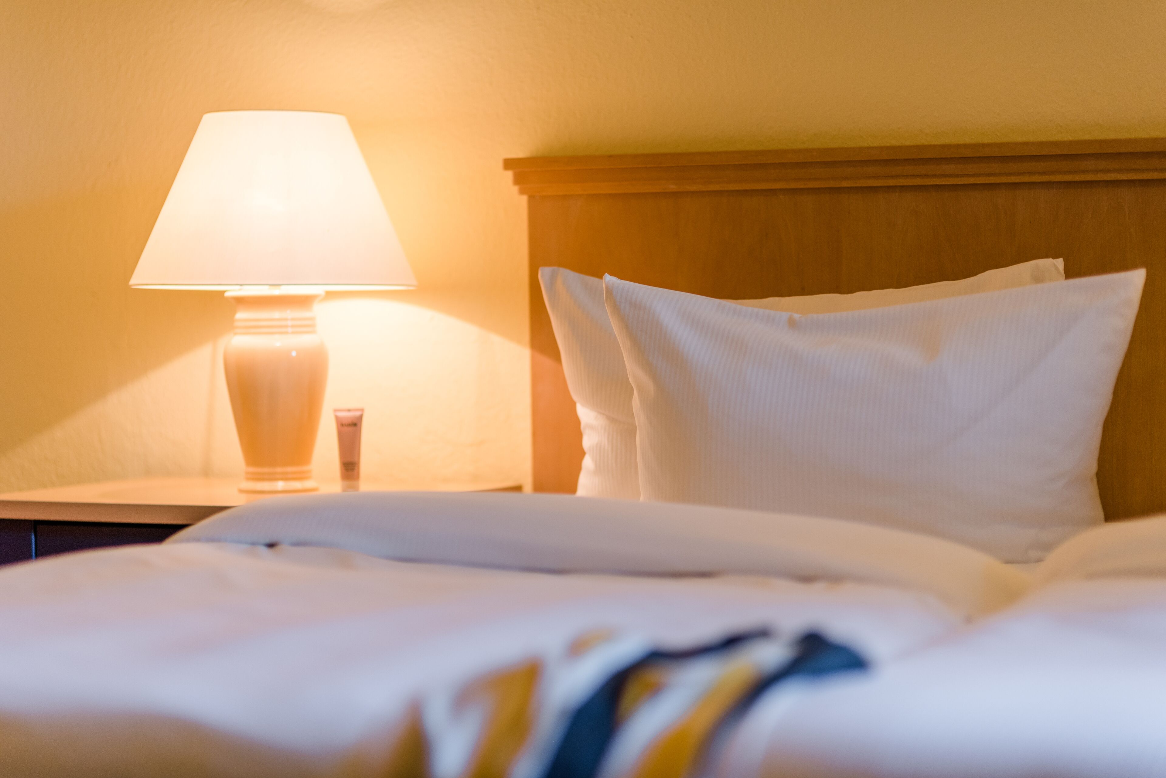 Una cama bien hecha con ropa blanca y almohadas, una lámpara de noche y un cabecero de madera.