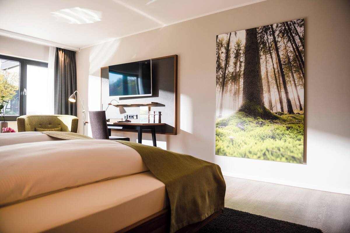A modern hotel room with a large bed, a desk with a computer, a TV, and a large forest-themed painting on the wall.