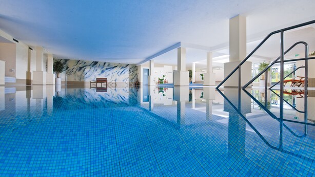 An indoor swimming pool with clear blue water, surrounded by white pillars and a modern design. - Sporthotel Grafenwald (Daun)