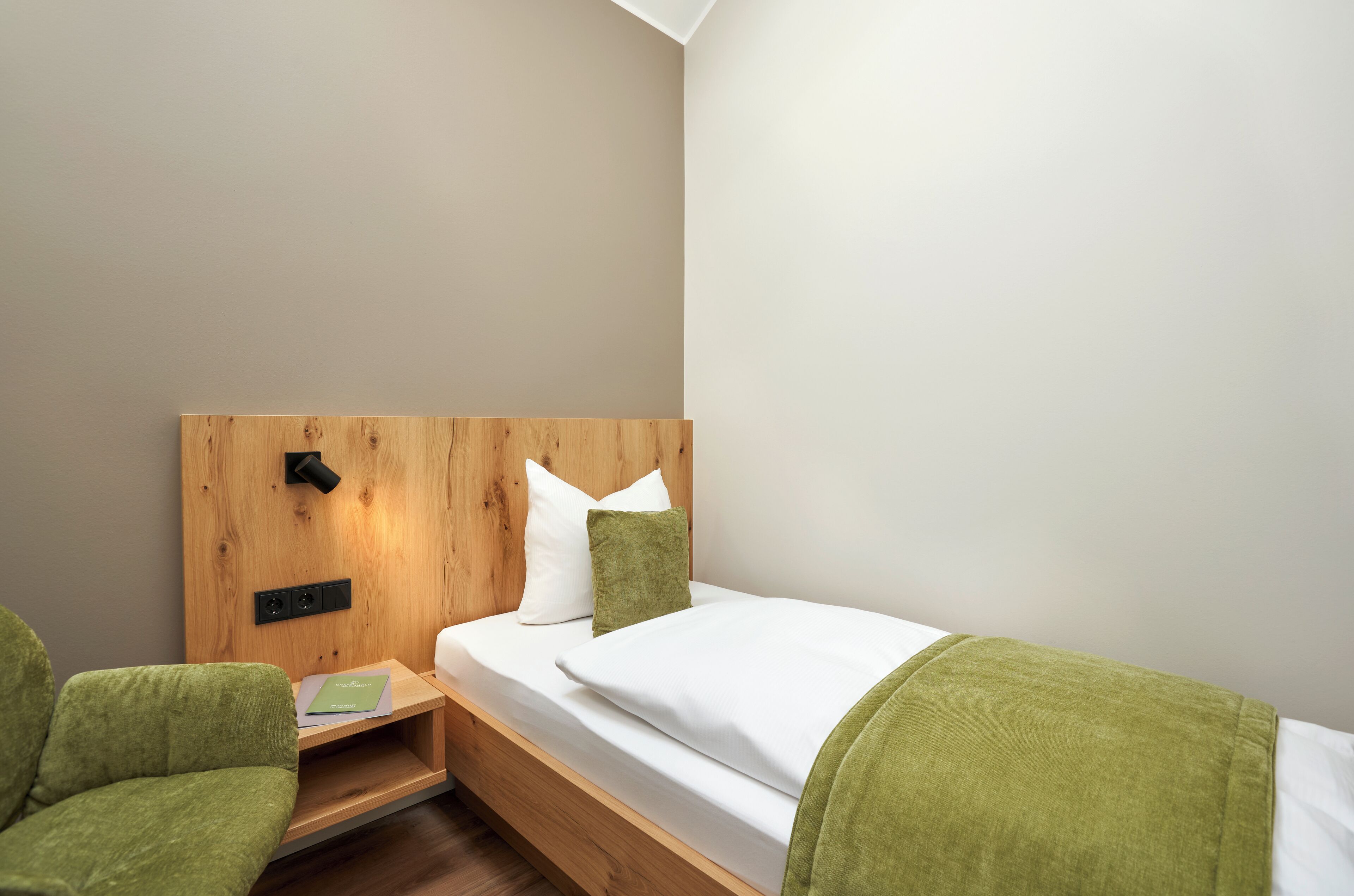 Une petite chambre à coucher, bien rangée, avec une tête de lit en bois, un lit simple avec une literie blanche, un plaid vert et une chaise verte.