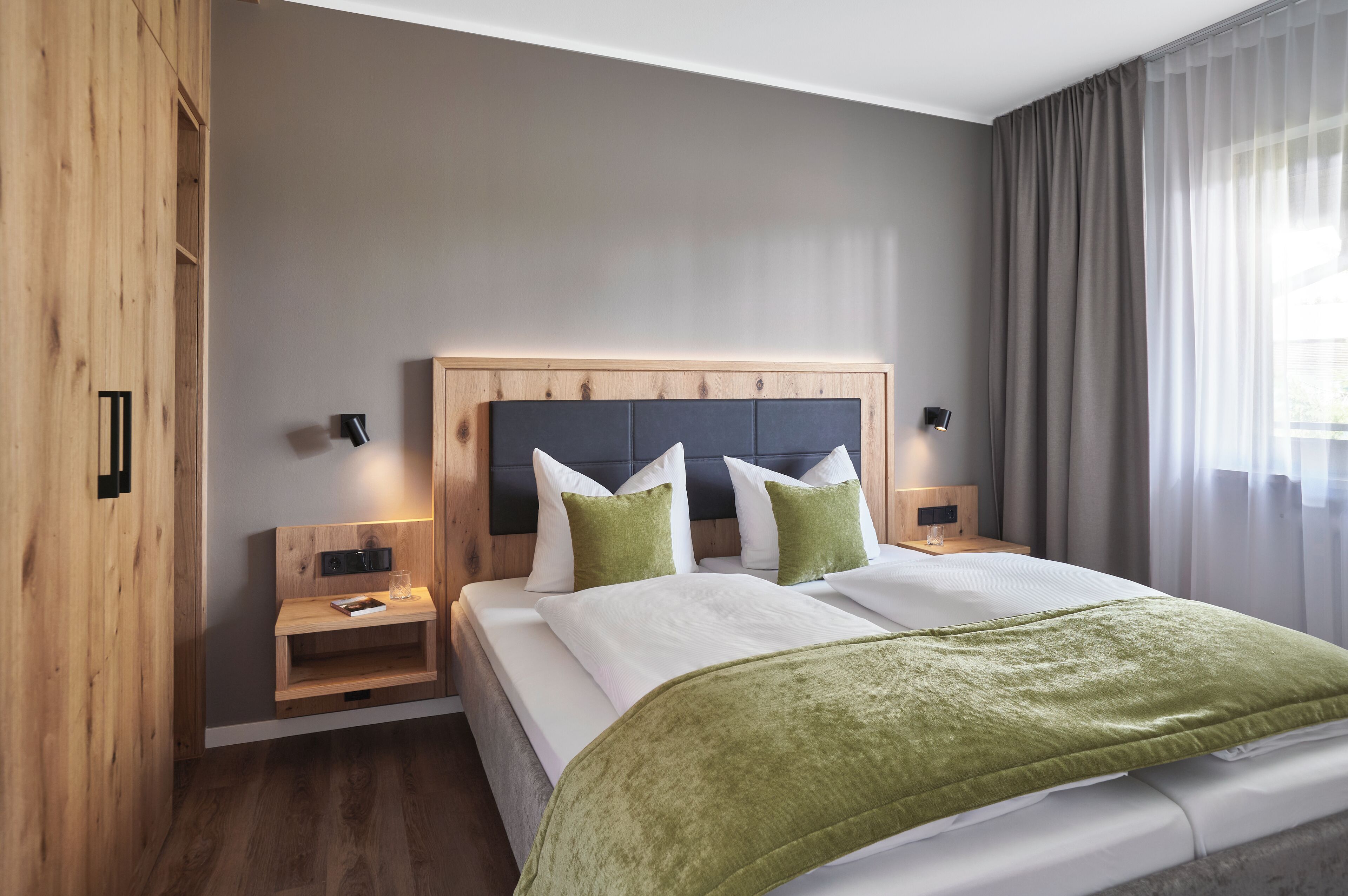 A modern hotel room with a wooden headboard, a green throw blanket, and a bedside table with a lamp.