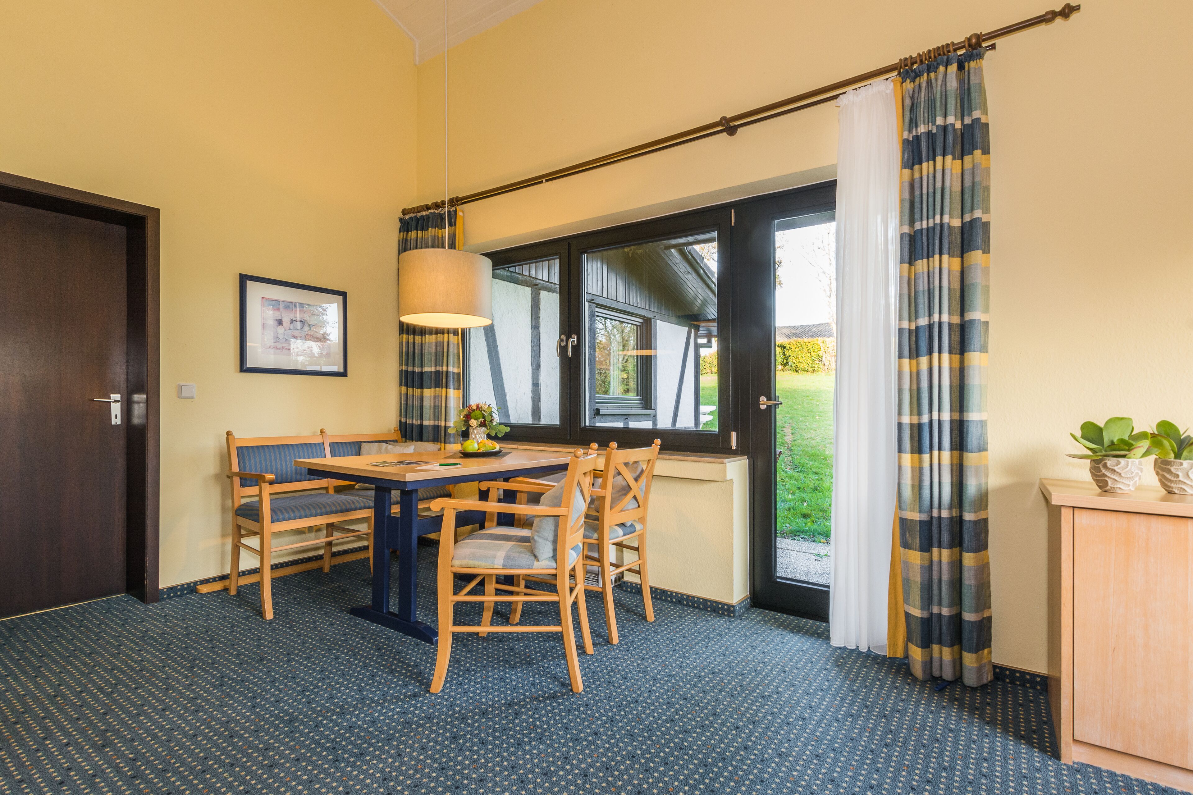 A room with a dining table and chairs, a lamp, a framed picture on the wall, and a window with curtains.