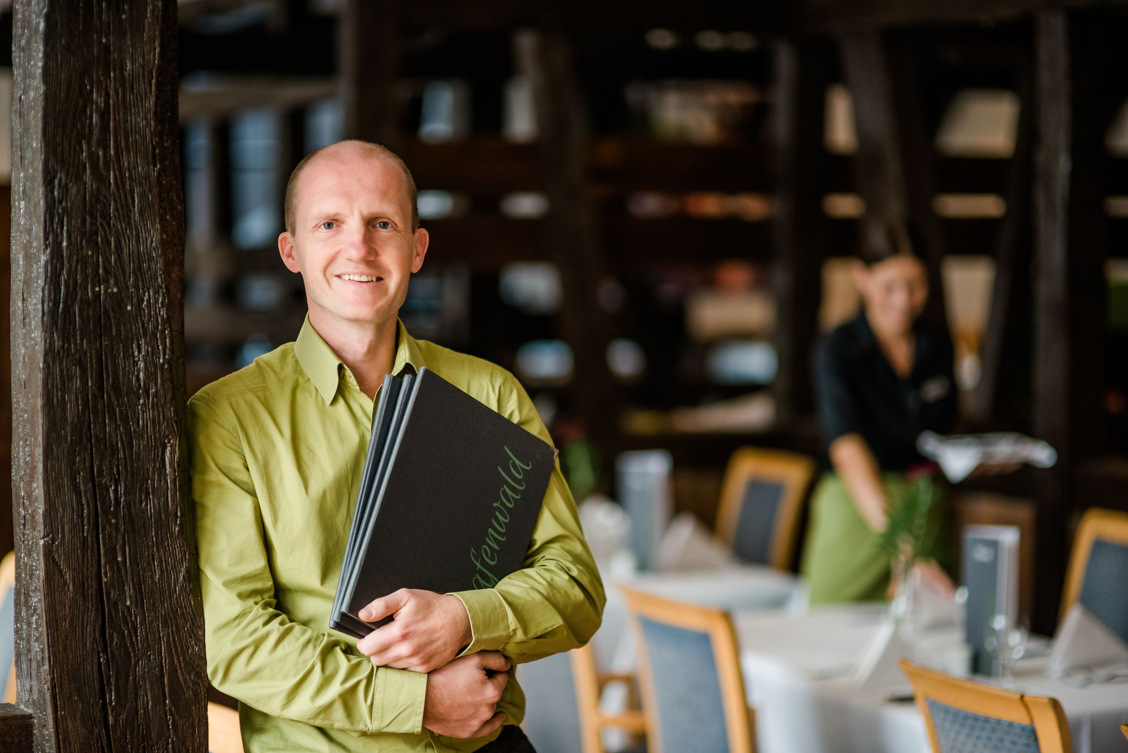 A person holding a black folder with 'afewald' written on it, standing in a restaurant with wooden beams and tables.