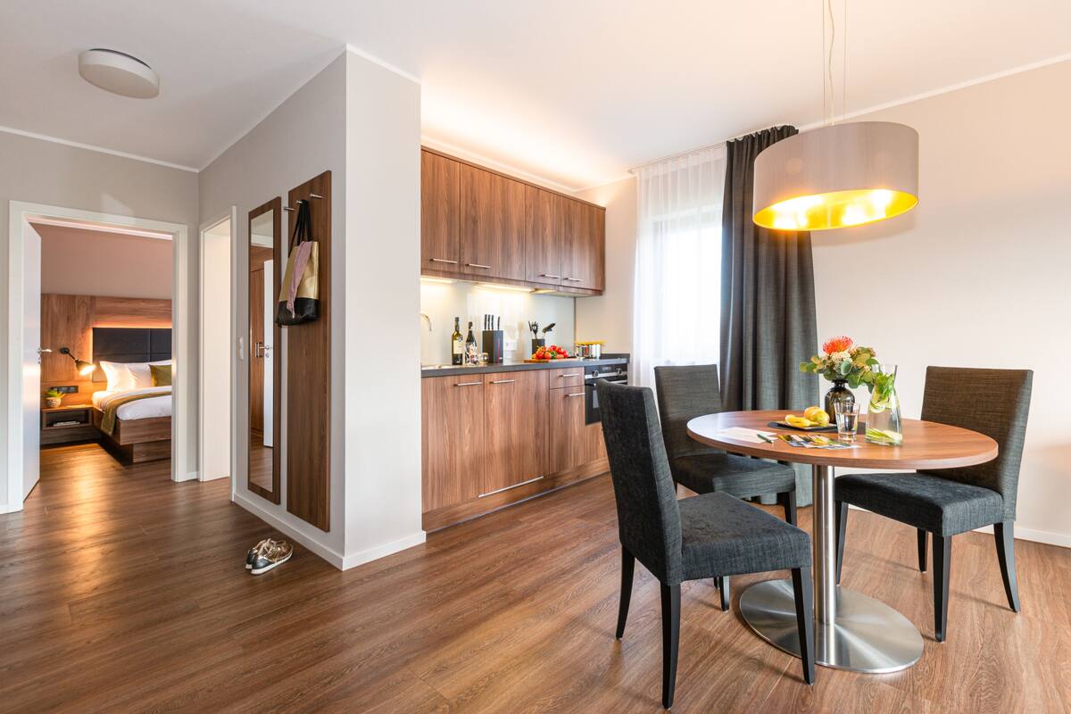 A modern hotel room with a wooden floor, a dining area with a round table and four chairs, a kitchen with wooden cabinets, and a large window with curtains.