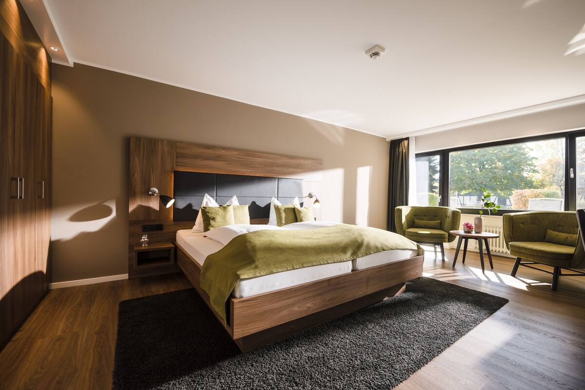 A modern bedroom with a large bed, a nightstand, a desk, two chairs, and a view of the outdoors.