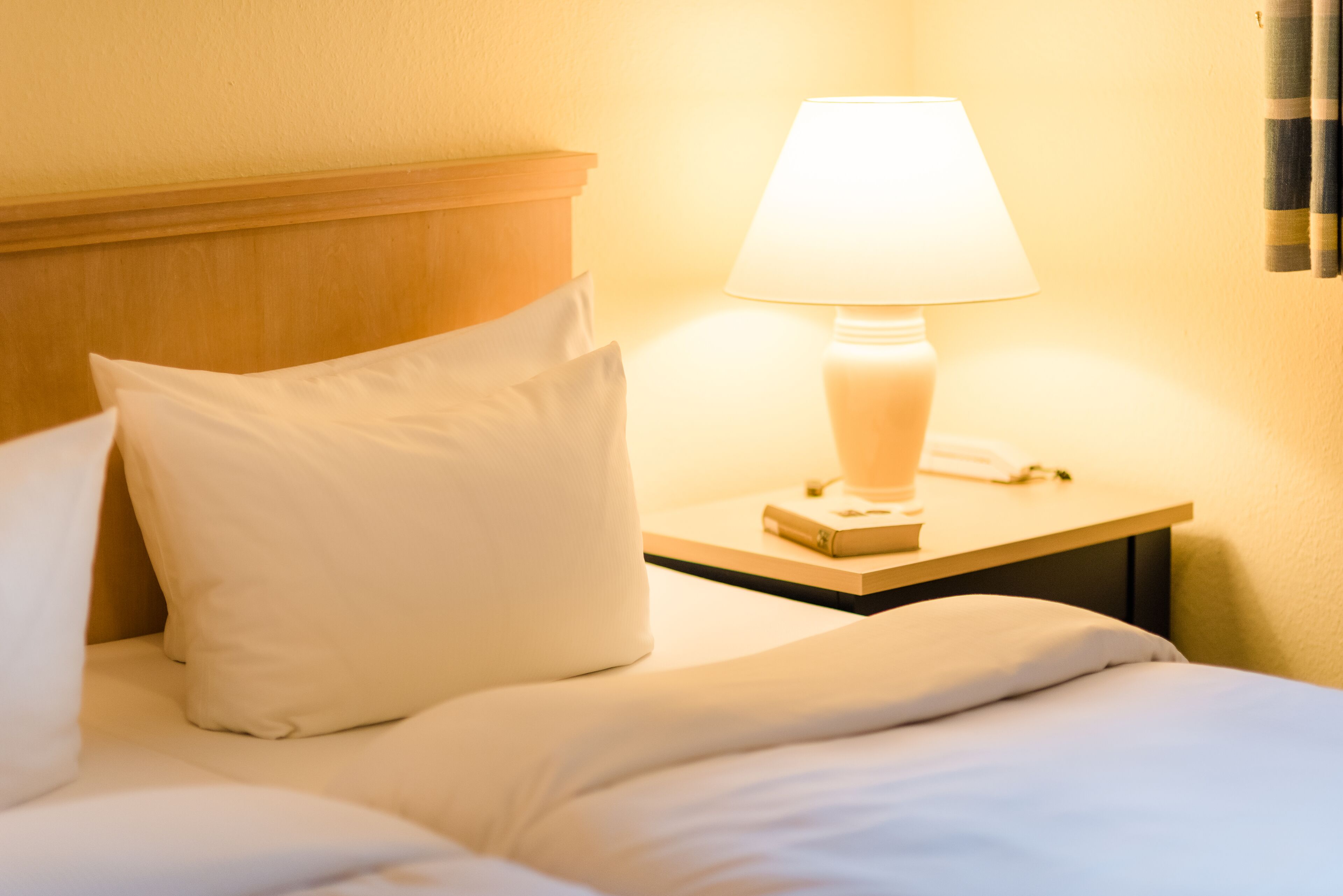 A neatly made bed with white bedding and pillows, a bedside table with a lamp, and a book.