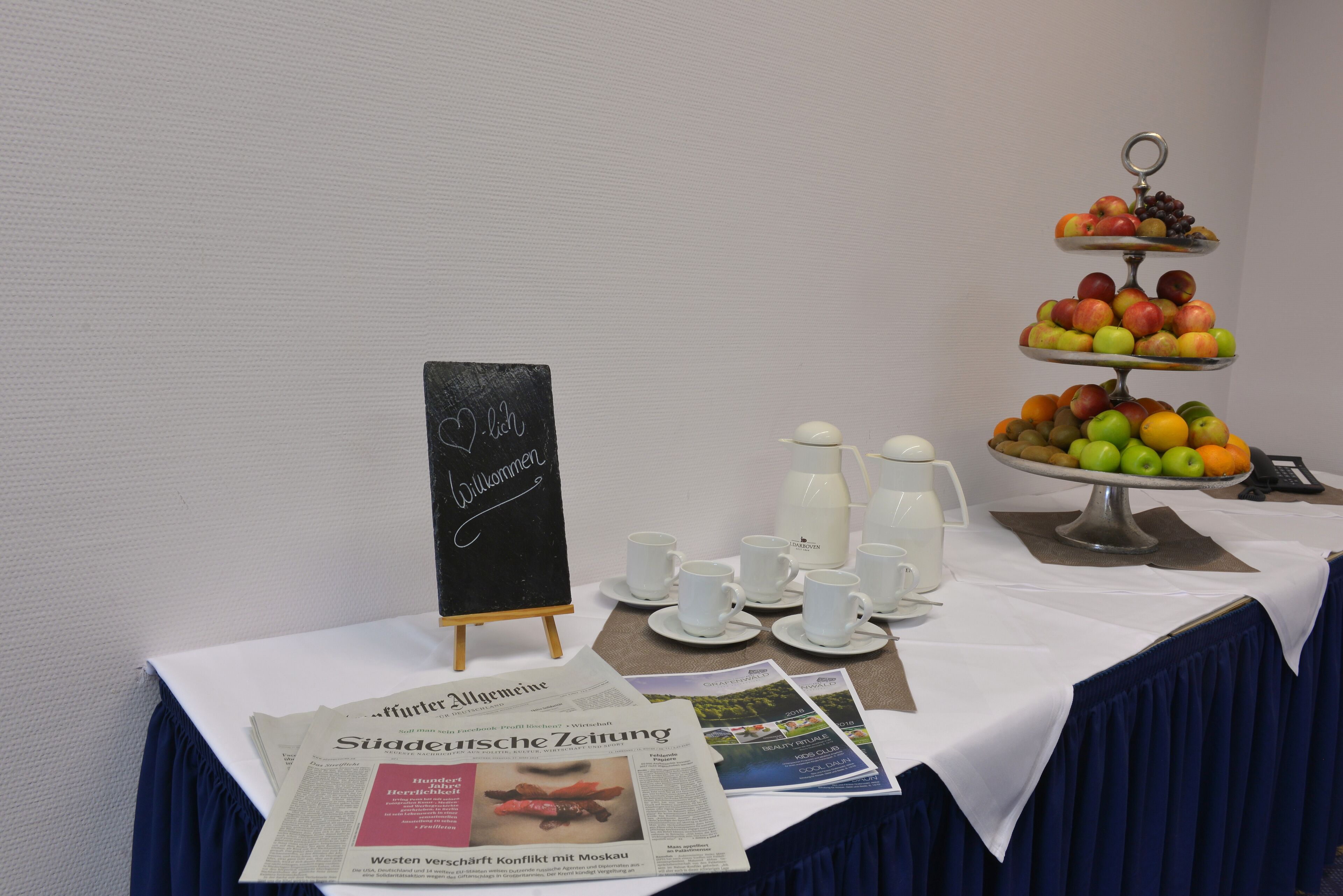 A table with a tiered fruit display, coffee cups, a milk pitcher, and newspapers.